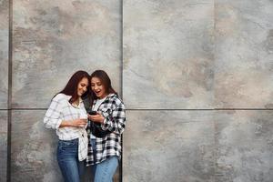 tenant une boisson et un téléphone. belles amies gaies ou couple de lesbiennes ensemble près du mur à l'extérieur pendant la journée photo