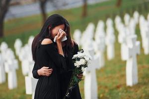 s'embrassent et pleurent. deux jeunes femmes en vêtements noirs visitant un cimetière avec de nombreuses croix blanches. conception des funérailles et de la mort photo