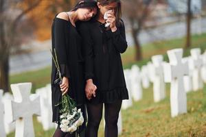 détient des fleurs. deux jeunes femmes en vêtements noirs visitant un cimetière avec de nombreuses croix blanches. conception des funérailles et de la mort photo