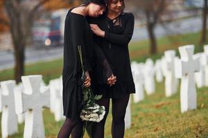 s'embrassent et pleurent. deux jeunes femmes en vêtements noirs visitant un cimetière avec de nombreuses croix blanches. conception des funérailles et de la mort photo