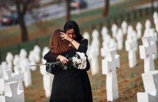 s'embrassent et pleurent. deux jeunes femmes en vêtements noirs visitant un cimetière avec de nombreuses croix blanches. conception des funérailles et de la mort photo