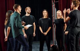 debout contre des rideaux rouges. groupe d'acteurs vêtus de vêtements de couleur sombre en répétition au théâtre photo