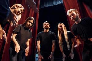 debout contre des rideaux rouges. groupe d'acteurs vêtus de vêtements de couleur sombre en répétition au théâtre photo