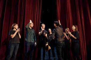 debout contre des rideaux rouges. groupe d'acteurs vêtus de vêtements de couleur sombre en répétition au théâtre photo