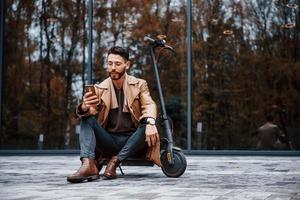 est assis sur un scooter électrique. jeune mannequin en vêtements à la mode est à l'extérieur de la ville pendant la journée photo