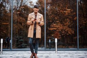 tient le smartphone. jeune mannequin en vêtements à la mode est à l'extérieur de la ville pendant la journée photo