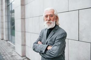 homme d'affaires senior en vêtements formels, avec des cheveux gris et une barbe est à l'extérieur se tient contre le mur photo