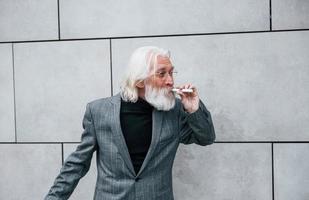 fumer une cigarette électronique. homme d'affaires senior en vêtements formels, aux cheveux gris et à la barbe est à l'extérieur photo