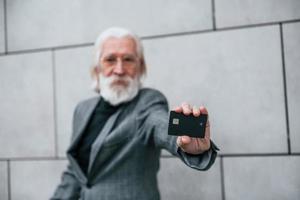 homme d'affaires senior en vêtements formels, aux cheveux gris et à la barbe est à l'extérieur détient une carte de crédit photo