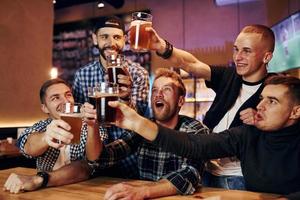 acclamant en frappant des verres à bière. groupe de personnes ensemble à l'intérieur dans le pub s'amuser le week-end photo