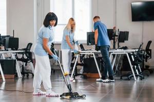 femme utilise un aspirateur. un groupe de travailleurs nettoie un bureau moderne ensemble pendant la journée photo