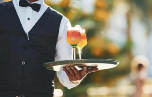 tient des cocktails. serveur noir en vêtements formels est à son travail à l'extérieur pendant la journée ensoleillée photo