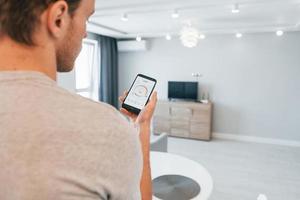 tient le téléphone. vue arrière d'un homme adulte qui contrôle à l'intérieur la technologie de la maison intelligente photo