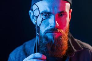 avec des ciseaux. jeune barbier barbu en casquette debout dans le studio avec éclairage au néon photo