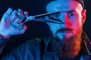 avec des ciseaux. jeune barbier barbu en casquette debout dans le studio avec éclairage au néon photo