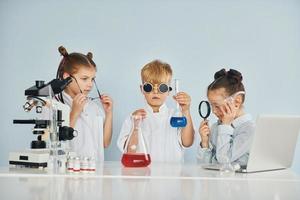 debout près de la table avec des tubes à essai. des enfants en blouse blanche jouent un scientifique en laboratoire en utilisant un équipement photo