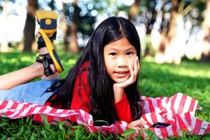 fille asiatique allongée avec ses mains sur son menton jouant au téléphone portable sur la pelouse du parc. concept, enfants jouant à l'extérieur, portrait d'enfant. mise au point douce et sélective. photo