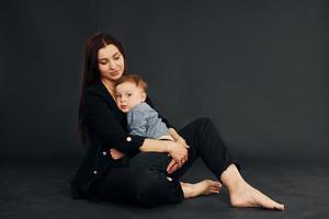 assis sur le sol. mère en vêtements noirs élégants est avec son petit fils dans le studio photo