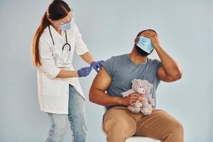 mec effrayé avec ours en peluche. femme médecin donnant une injection à un homme afro-américain à l'hôpital photo