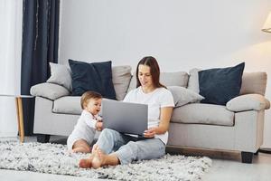 à l'aide d'un ordinateur portable. mère avec sa petite fille est à l'intérieur à la maison ensemble photo