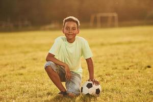 est assis avec un ballon de football. un enfant afro-américain s'amuse sur le terrain pendant la journée d'été photo