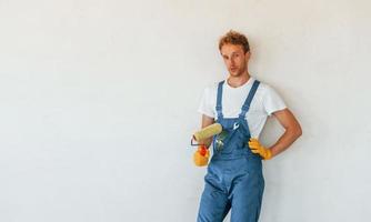 debout contre des murs blancs finis. jeune homme travaillant en uniforme à la construction pendant la journée photo