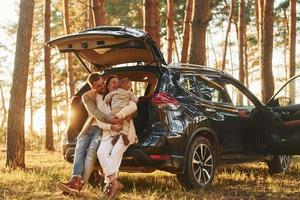 appuyé sur la voiture. famille heureuse de père, mère et petite fille est dans la forêt photo