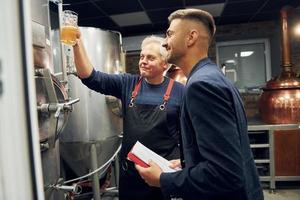 tenant du verre. un homme âgé et un jeune travailleur avec un document se tiennent dans la salle de stockage de la bière photo