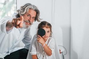 ophtalmologiste masculin senior aux cheveux gris et à la barbe en blouse blanche est à l'intérieur dans une clinique testant la vue d'une petite fille photo