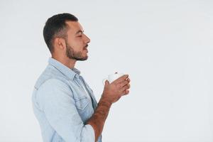 tient une tasse blanche. jeune bel homme debout à l'intérieur sur fond blanc photo