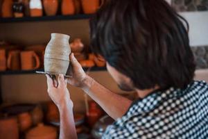 le jeune céramiste tient un pot fait main frais à la main et examine les résultats de son travail photo