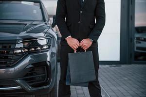 vue rapprochée d'un homme d'affaires en costume noir et cravate se tient près d'une automobile moderne avec des sacs à provisions dans les mains photo