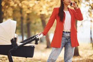 parler au téléphone. mère en manteau rouge se promener avec son enfant dans le landau dans le parc à l'automne photo