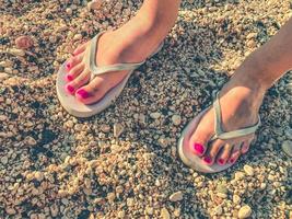 une fille avec une pédicure rose aux pieds se promène dans un pays chaud dans des pantoufles en caoutchouc blanc. chaussures de loisirs se dresse sur le sable. ardoises pour se promener le long de la côte de la mer photo