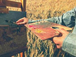 l'artiste peint sur toile. entre les mains de l'artiste un pinceau et une palette de couleurs. mélangé plusieurs couleurs pour peindre un tableau. l'homme dessine sur le fond d'un champ jaune photo