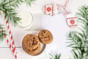 vue de dessus à travers les branches de sapin vert au verre de lait, pailles, biscuits, page vide, mitaines sur tableau blanc photo