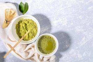 tasse à thé vert matcha et bol à poudre, cuillère en bois, fouet en bambou, feuilles fraîches sur une serviette sur fond texturé gris. photo