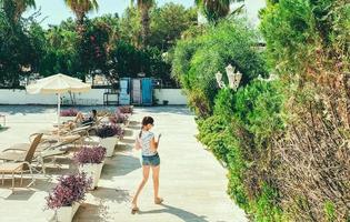 une fille avec une coiffure en short et un t-shirt se promène dans l'hôtel près de la piscine. chaises longues à proximité pour les touristes, buissons verts et plantes photo