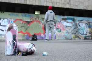 plusieurs bombes aérosols usagées avec de la peinture rose et blanche se trouvent sur l'asphalte contre le gars debout devant un mur peint dans des dessins de graffitis colorés photo