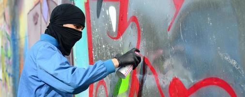 un jeune hooligan au visage caché peint des graffitis sur un mur métallique. concept de vandalisme illégal photo