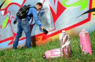 quelques pots de peinture usagés sur le fond de l'espace avec le mur sur lequel le jeune homme dessine un grand dessin graffiti. art moderne de dessiner des murs en graffiti photo