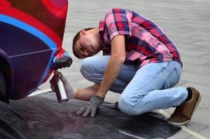 un jeune graffeur aux cheveux roux peint un nouveau graffiti coloré sur la voiture. photo du processus de dessin d'un graffiti sur un gros plan de voiture. le concept d'art de rue et de vandalisme illégal