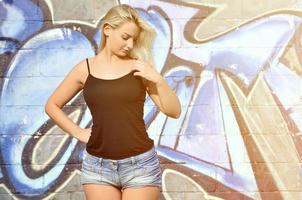 fille blonde caucasienne sexy en short en jean et débardeur noir posant contre un mur de graffitis pendant la journée à l'extérieur photo