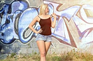 fille blonde caucasienne sexy en short en jean et débardeur noir posant contre un mur de graffitis pendant la journée à l'extérieur photo