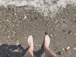 plage de galets avec des pierres. un gars en chaussures se tient sur le rivage. reste de touristes sur la mer. les pieds lavés par les vagues de la mer photo