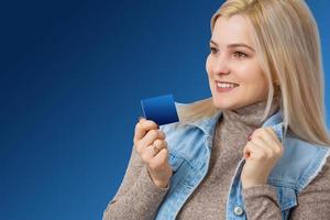 portrait d'une jolie jeune femme blonde en chandail montrant une carte de crédit et regardant loin l'espace de copie isolé sur fond bleu photo