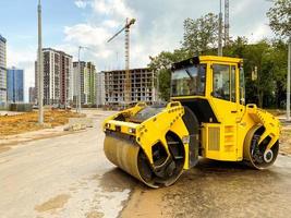 machines de construction construit une grande zone résidentielle. pose des communications. un finisseur d'asphalte jaune vif compacte l'asphalte sur la route sur fond de grands immeubles résidentiels photo