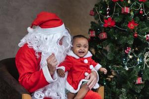 le père noël soulève une petite fille heureuse en bas âge et rit joyeusement avec un arbre de noël entièrement décoré à l'arrière pour le concept de célébration de la saison photo