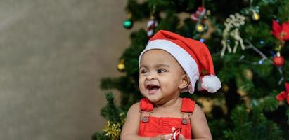 bébé afro-américain en bas âge sourit joyeusement tout en s'habillant dans des vêtements de noël et un bonnet de noel avec un arbre de noël sur le dos pour le concept de célébration de la saison photo