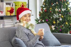 senior caucasian woman sitting on the sofa couch wearing red santa hat tout en buvant du chocolat chaud avec l'arbre de noël sur le dos photo